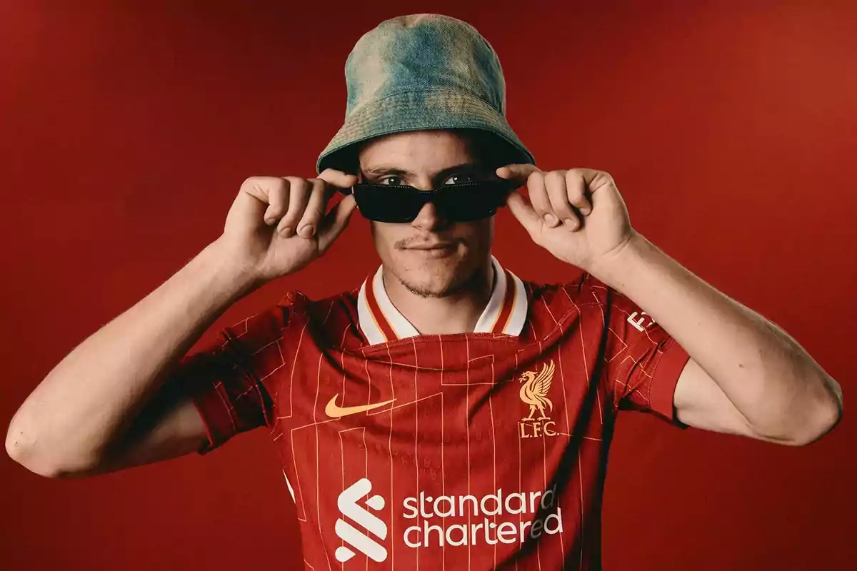 Person wearing a Liverpool jersey, denim hat, and dark sunglasses against a red background Person wearing a Liverpool jersey, denim hat, and dark sunglasses against a red background