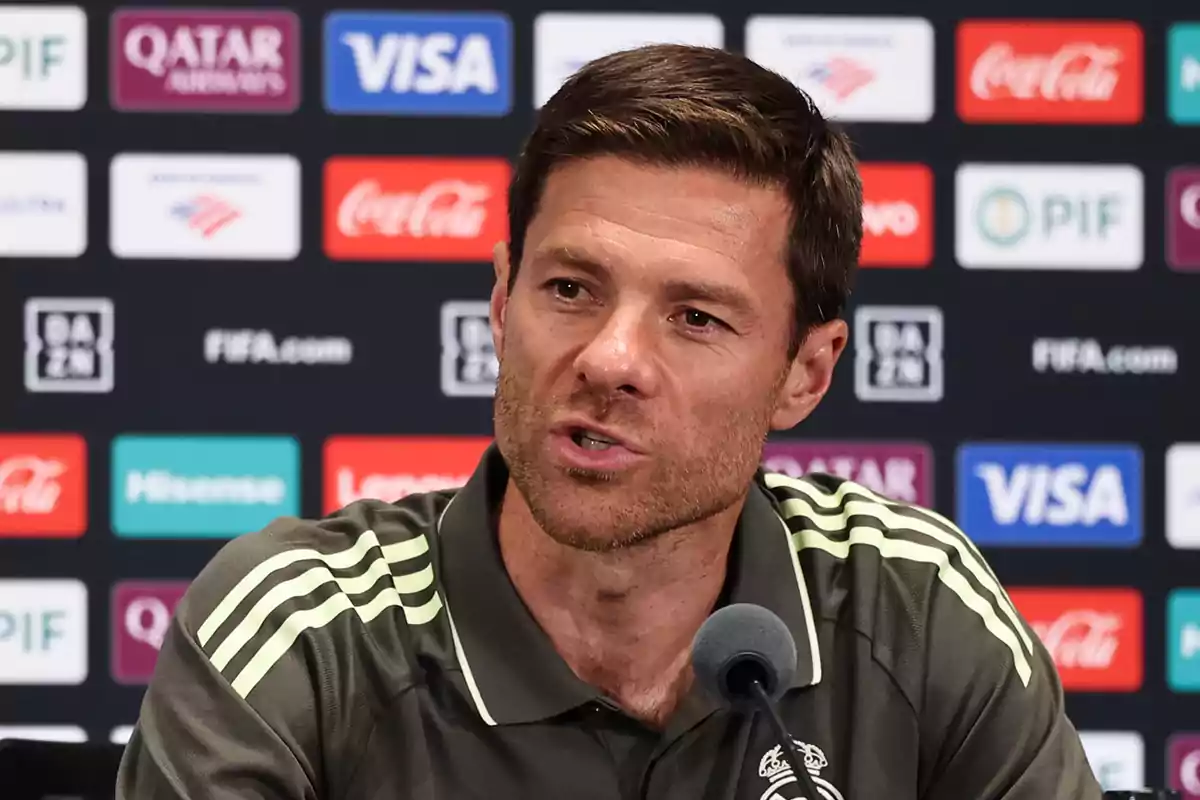 Hombre con camiseta deportiva hablando frente a un micrófono en una rueda de prensa con fondo de logotipos de patrocinadores. Hombre con camiseta deportiva hablando frente a un micrófono en una rueda de prensa con fondo de logotipos de patrocinadores.