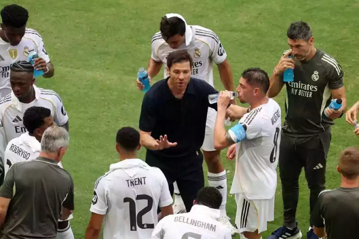 Un entrenador de fútbol da indicaciones a sus jugadores durante una pausa en el partido mientras varios futbolistas beben agua en el campo