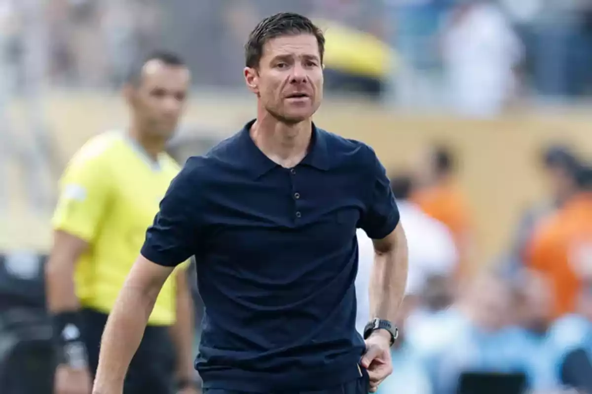 Hombre con camiseta azul marino en un campo de fútbol con un árbitro al fondo y personas desenfocadas en las gradas