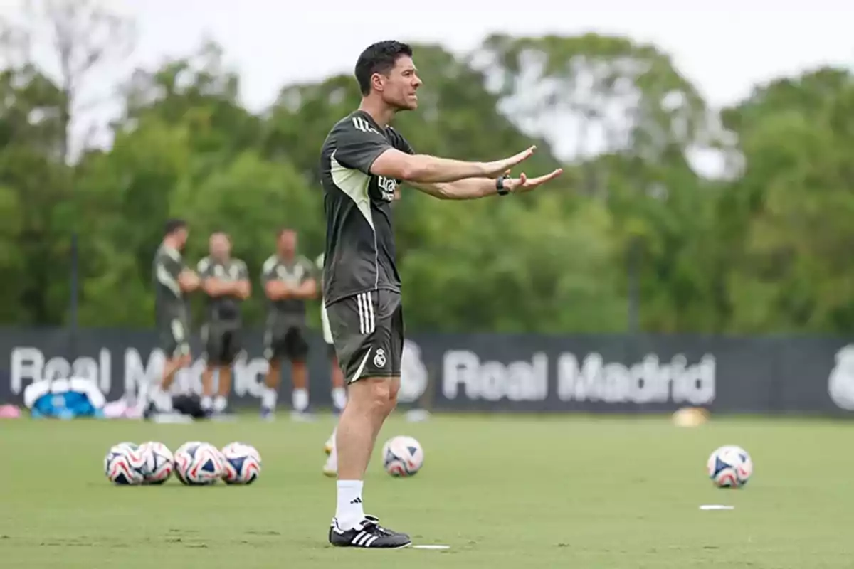 Entrenador de fútbol dando indicaciones en un campo de entrenamiento con balones y jugadores al fondo Entrenador de fútbol dando indicaciones en un campo de entrenamiento con balones y jugadores al fondo
