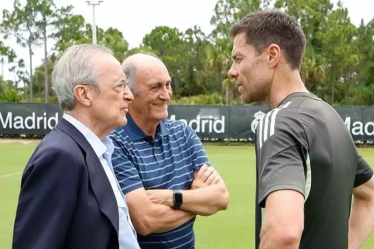 Tres hombres conversan en un campo de entrenamiento de fútbol con árboles y un cartel de Madrid al fondo