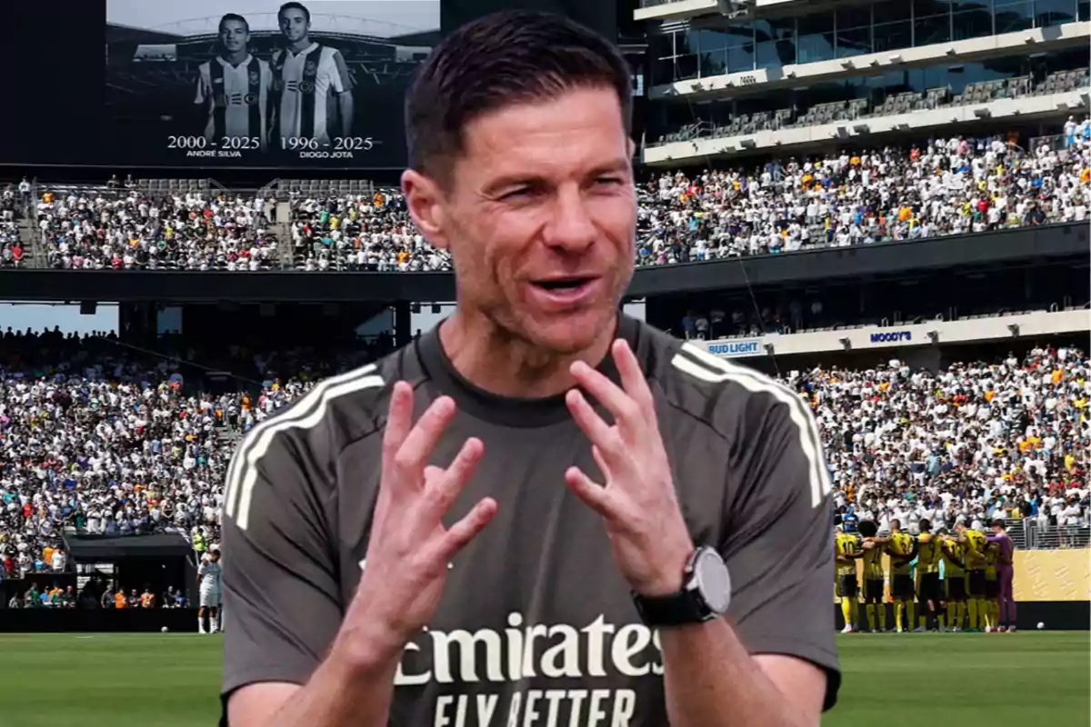 Entrenador de fútbol con camiseta de entrenamiento gesticulando con las manos en un estadio lleno de aficionados y un grupo de jugadores reunidos al fondo