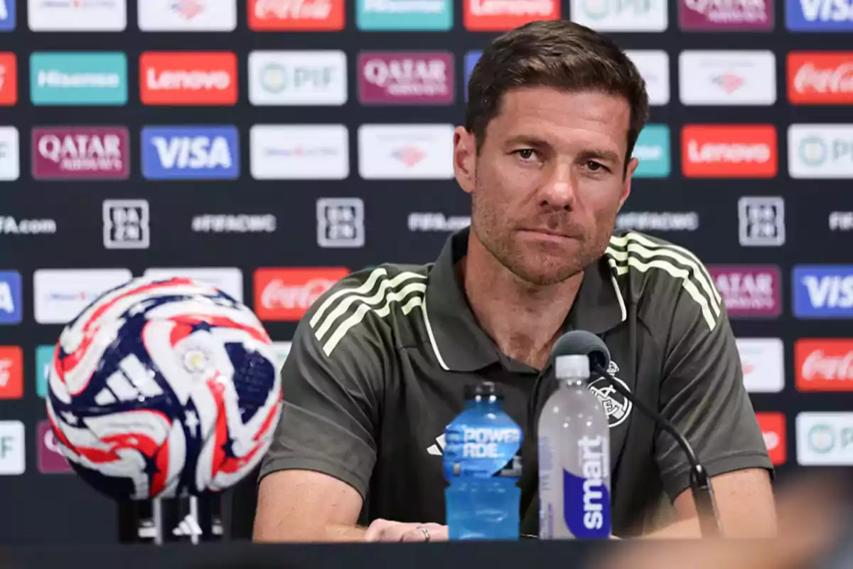 Soccer coach sitting at a press conference with a ball, water bottles, and a microphone in front of him, background with sponsor logos.