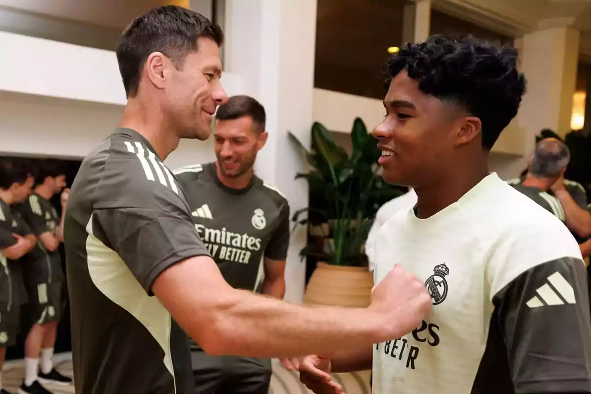 Entrenador y jugador del Real Madrid sonríen y se saludan durante un entrenamiento en interiores, ambos llevan uniforme deportivo del equipo