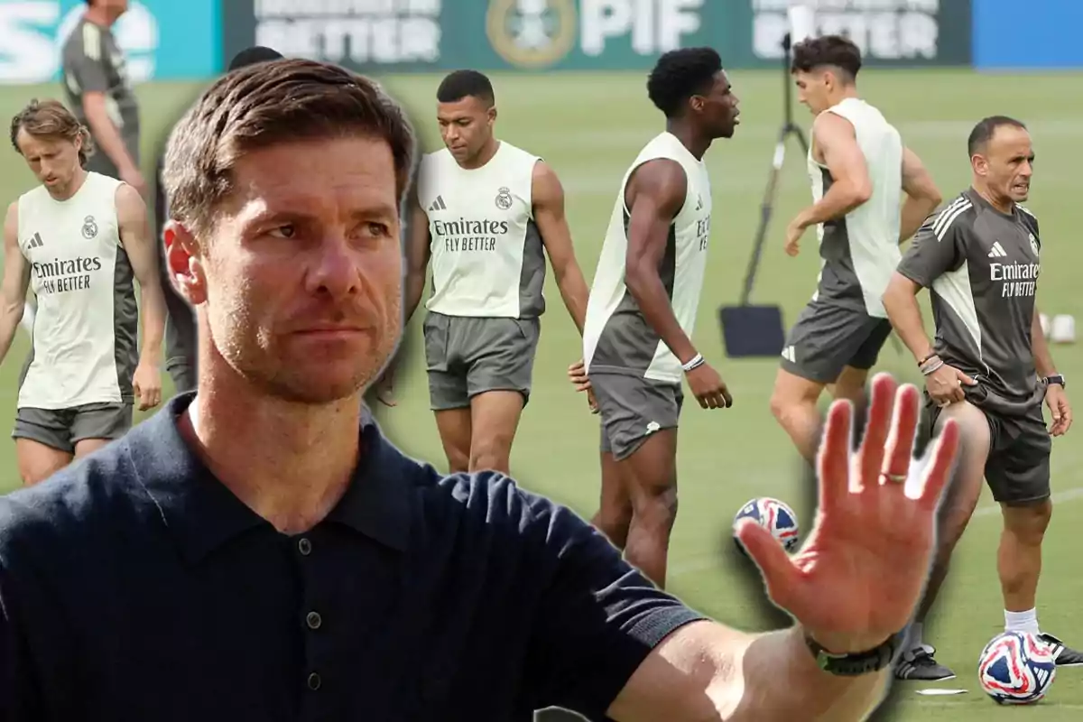 Un hombre en primer plano con expresión seria y la mano levantada mientras al fondo varios jugadores de fútbol entrenan en un campo con uniformes del Real Madrid