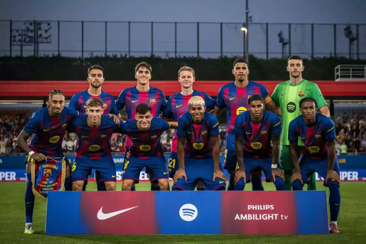 Once jugadores del FC Barcelona posan juntos en el campo antes de un partido, todos llevan el uniforme azul y grana y están detrás de un cartel con los logotipos de Nike, Spotify y Philips Ambilight TV. Once jugadores del FC Barcelona posan juntos en el campo antes de un partido, todos llevan el uniforme azul y grana y están detrás de un cartel con los logotipos de Nike, Spotify y Philips Ambilight TV.
