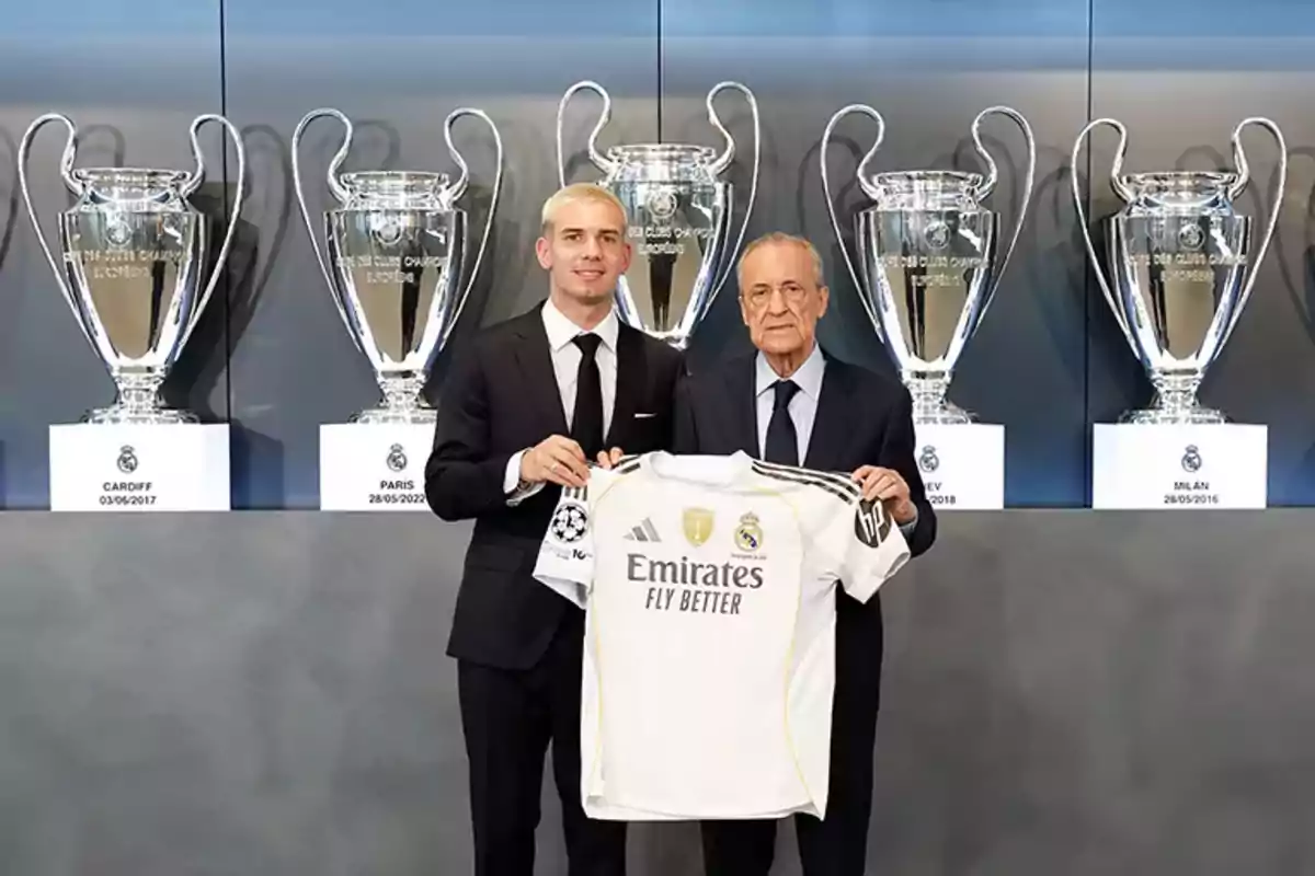 Dos hombres con traje posan sosteniendo una camiseta del Real Madrid frente a una vitrina con trofeos de la Champions League.