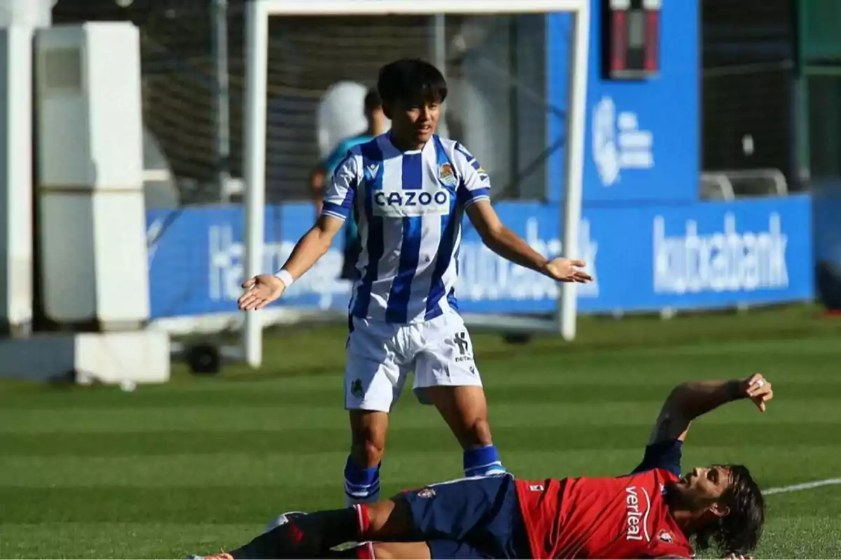 Un jugador de la Real Sociedad con los brazos abiertos mira a un rival de Osasuna que está tirado en el césped durante un partido de fútbol. Un jugador de la Real Sociedad con los brazos abiertos mira a un rival de Osasuna que está tirado en el césped durante un partido de fútbol.