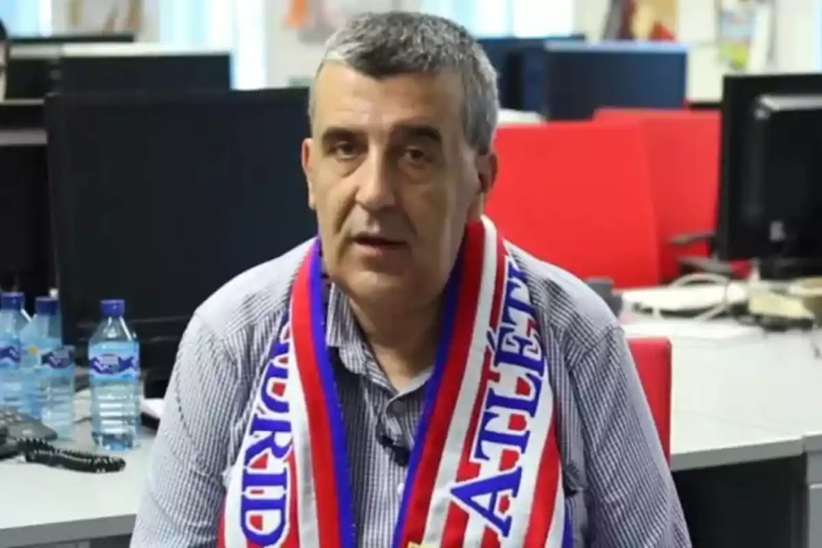 Gray-haired man with an Atlético de Madrid scarf sitting in an office with desks and water bottles in the background Gray-haired man with an Atlético de Madrid scarf sitting in an office with desks and water bottles in the background