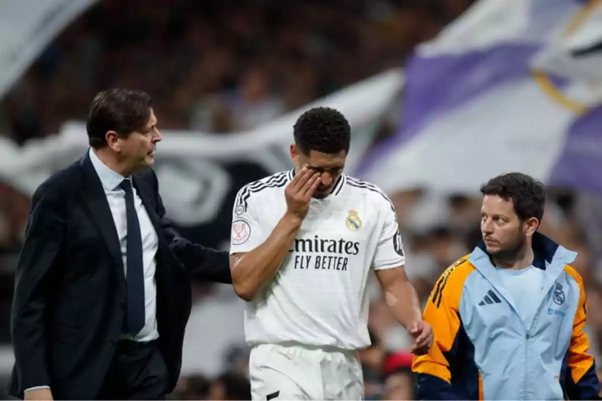 Un jugador del Real Madrid se retira del campo acompañado por dos miembros del staff mientras se cubre el rostro con la mano en un estadio lleno. Un jugador del Real Madrid se retira del campo acompañado por dos miembros del staff mientras se cubre el rostro con la mano en un estadio lleno.