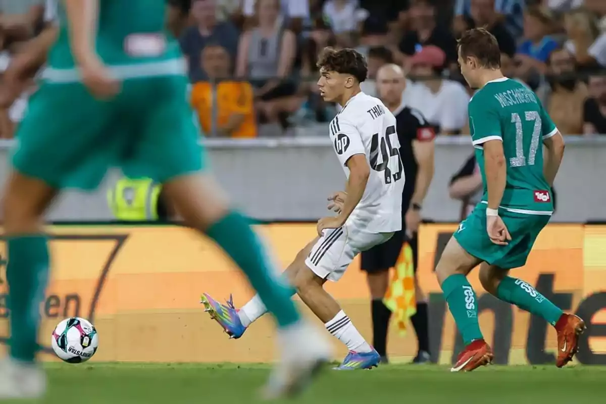 Un jugador de fútbol con uniforme blanco controla el balón mientras es presionado por dos rivales con uniforme verde durante un partido en un estadio lleno de espectadores. Un jugador de fútbol con uniforme blanco controla el balón mientras es presionado por dos rivales con uniforme verde durante un partido en un estadio lleno de espectadores.