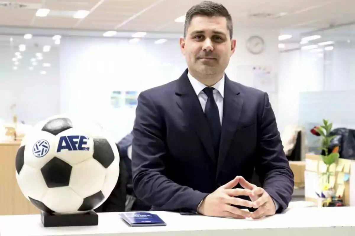 Hombre de traje oscuro posando en una oficina junto a un balón de fútbol blanco y negro con el logo de AFE Hombre de traje oscuro posando en una oficina junto a un balón de fútbol blanco y negro con el logo de AFE