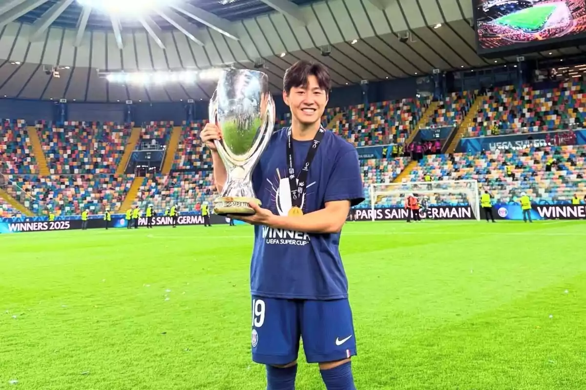 Futbolista sonriente sostiene un gran trofeo plateado en el campo de un estadio lleno de asientos coloridos tras ganar la Supercopa de la UEFA Futbolista sonriente sostiene un gran trofeo plateado en el campo de un estadio lleno de asientos coloridos tras ganar la Supercopa de la UEFA