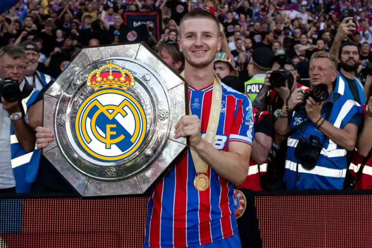Un futbolista sonriente sostiene un trofeo grande con el escudo del Real Madrid mientras fotógrafos y aficionados lo rodean en el estadio. Un futbolista sonriente sostiene un trofeo grande con el escudo del Real Madrid mientras fotógrafos y aficionados lo rodean en el estadio.