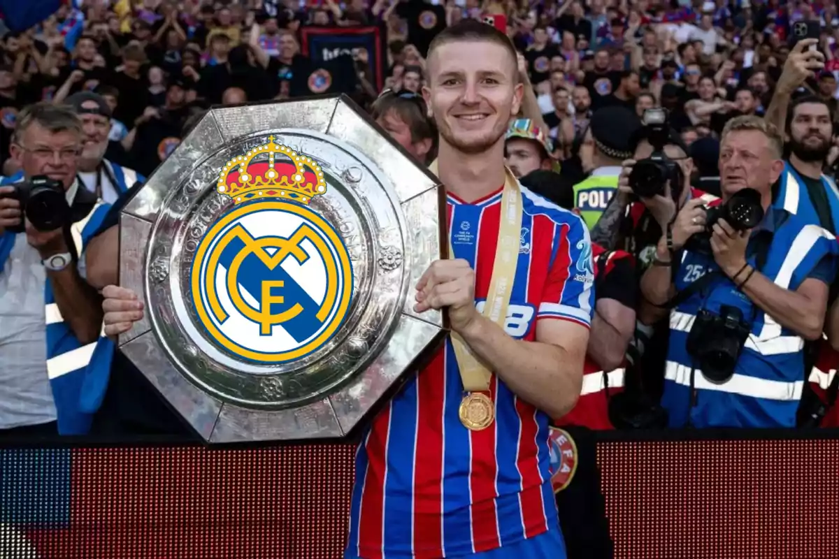 Un futbolista sonriente con uniforme de rayas rojas y azules sostiene un trofeo grande con el escudo del Real Madrid mientras fotógrafos y aficionados lo rodean en el estadio