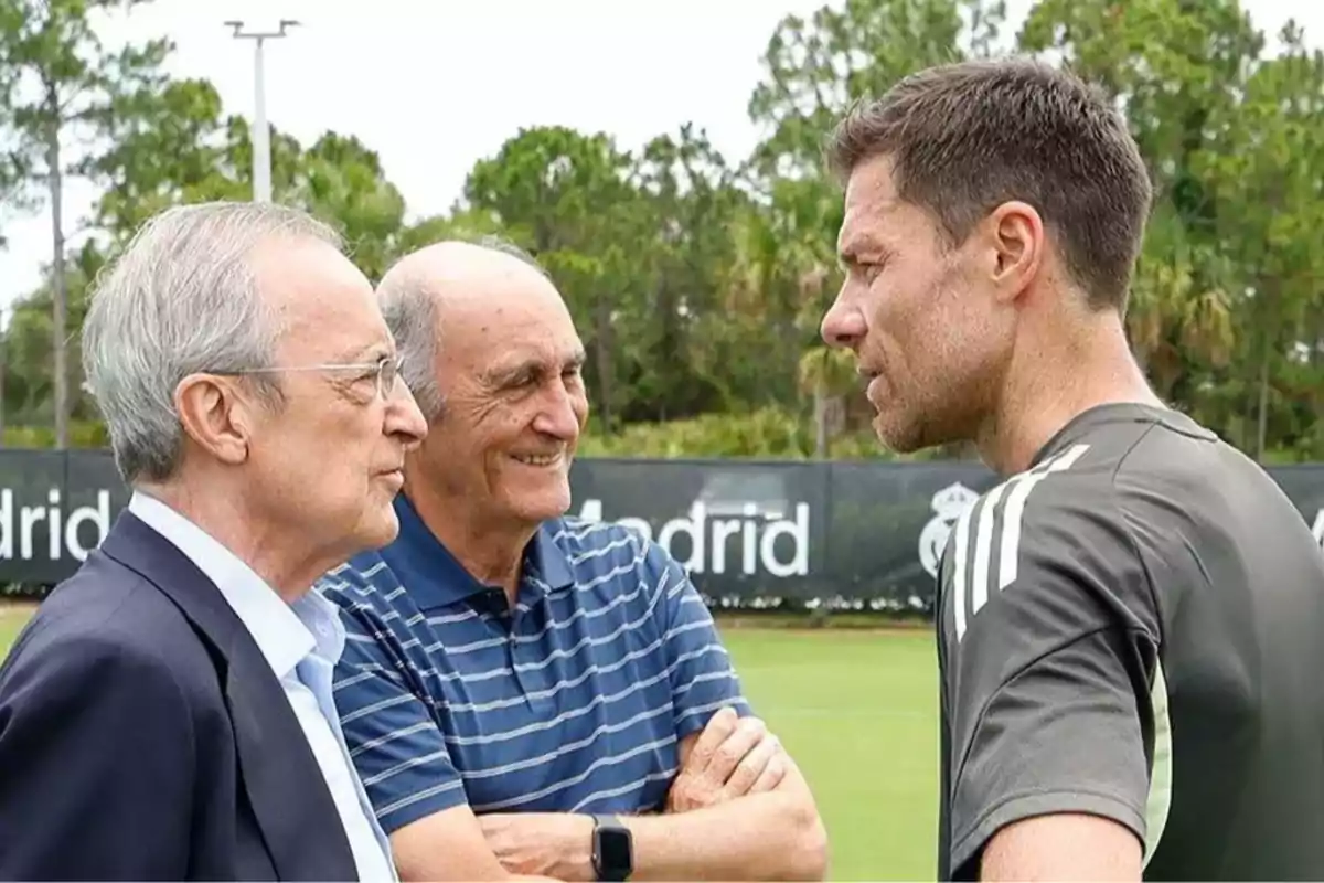Three men are talking on a soccer training field with trees in the background and a Madrid sign behind them. Three men are talking on a soccer training field with trees in the background and a Madrid sign behind them.