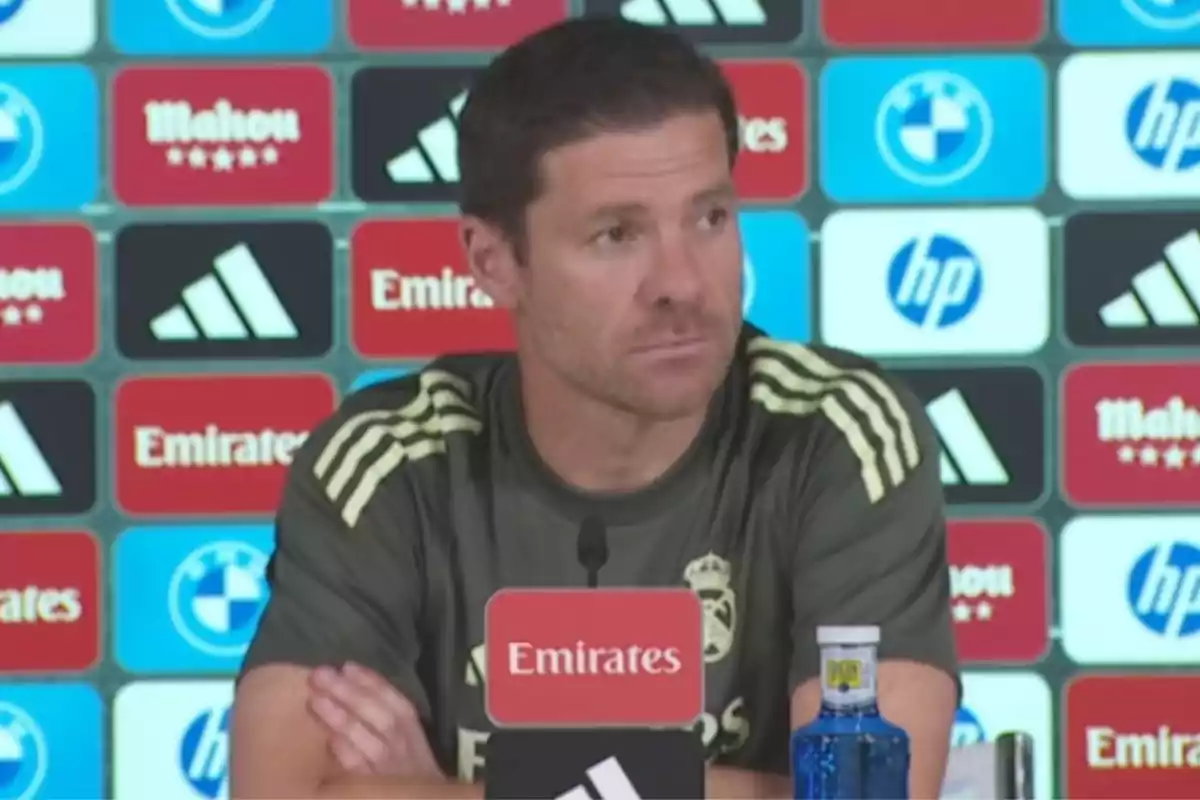Man wearing a Real Madrid training jersey sitting at a press conference in front of a microphone and a backdrop with sponsor logos