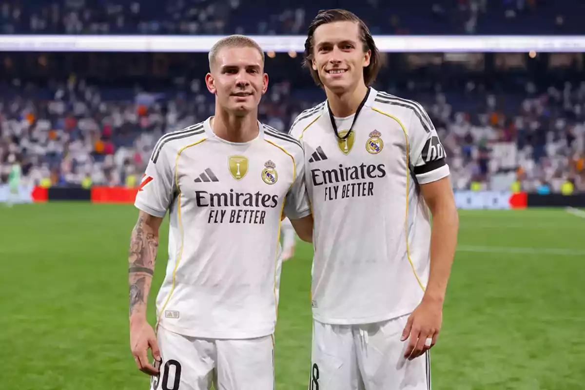 Two Real Madrid players pose smiling on the field after a match, wearing the team’s white uniform.