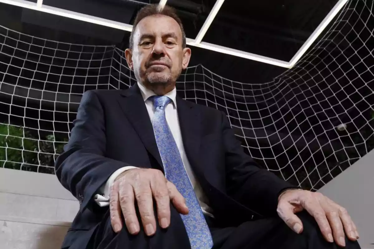 Hombre de traje oscuro y corbata azul sentado con expresión seria en un entorno moderno con una red blanca de fondo Hombre de traje oscuro y corbata azul sentado con expresión seria en un entorno moderno con una red blanca de fondo