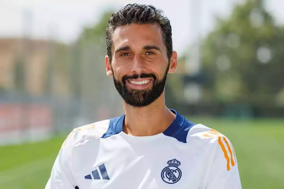 Hombre con barba y cabello oscuro sonríe mientras viste una camiseta blanca del Real Madrid en un campo de entrenamiento al aire libre Hombre con barba y cabello oscuro sonríe mientras viste una camiseta blanca del Real Madrid en un campo de entrenamiento al aire libre