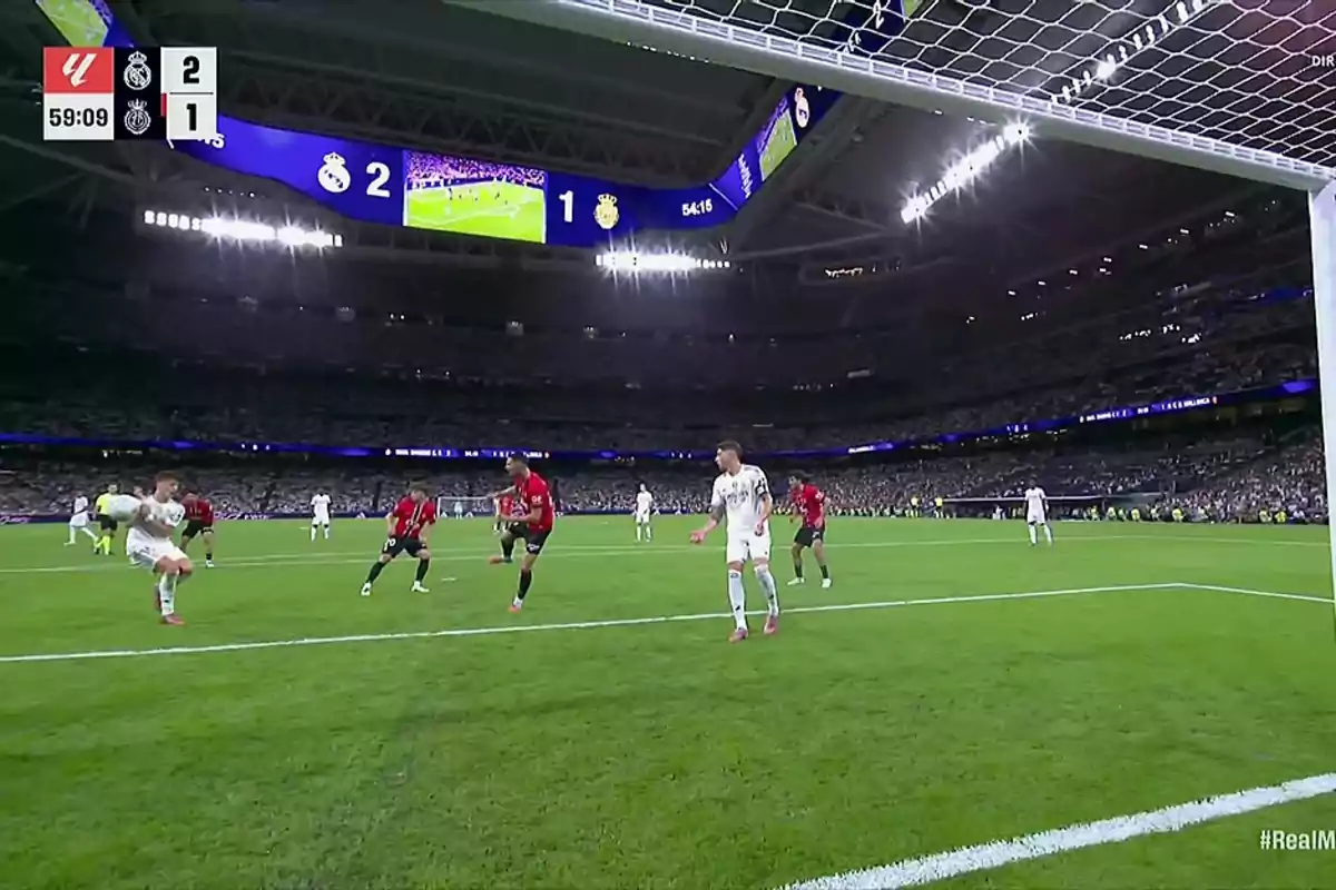 Un jugador de fútbol de uniforme blanco patea el balón cerca de la portería mientras varios jugadores con uniforme rojo intentan defender en un estadio lleno de espectadores. Un jugador de fútbol de uniforme blanco patea el balón cerca de la portería mientras varios jugadores con uniforme rojo intentan defender en un estadio lleno de espectadores.