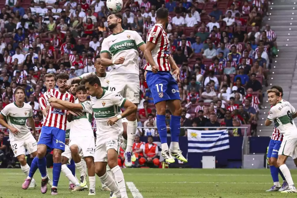 Jugadores de fútbol de dos equipos saltan para disputar un balón en el área durante un partido con el público observando desde las gradas. Jugadores de fútbol de dos equipos saltan para disputar un balón en el área durante un partido con el público observando desde las gradas.