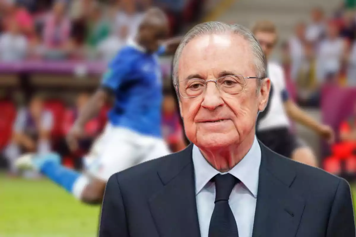 Older man with glasses and a dark suit in the foreground with a blurred soccer match in the background