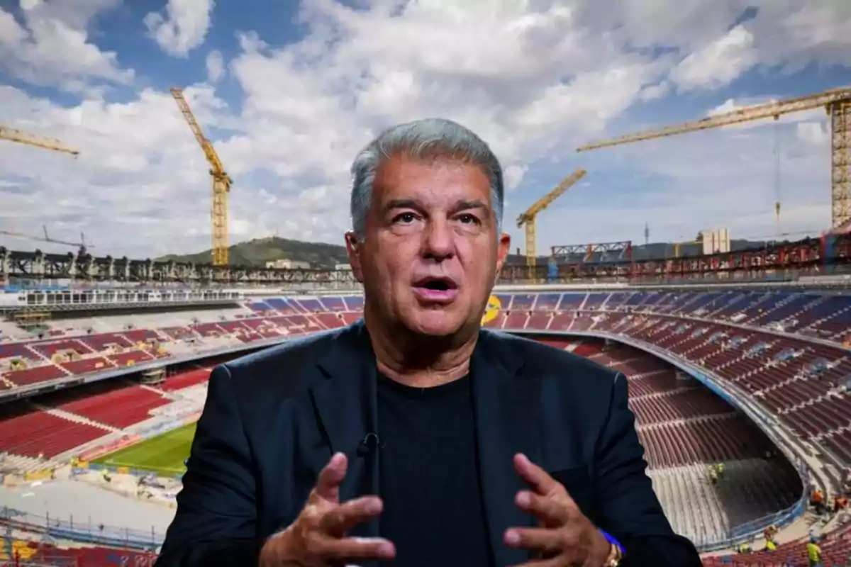 Gray-haired man in a dark suit speaking in front of a stadium under renovation with cranes and a partly cloudy sky in the background