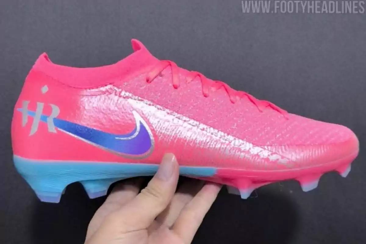 Pink Nike soccer cleat with blue details and a silver logo held by a hand against a dark background Pink Nike soccer cleat with blue details and a silver logo held by a hand against a dark background