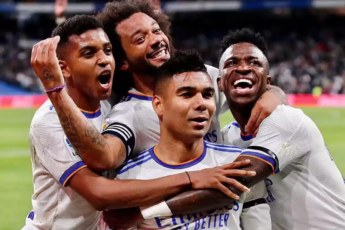 Cuatro futbolistas con uniforme blanco celebran abrazados y sonrientes en un estadio de fútbol
