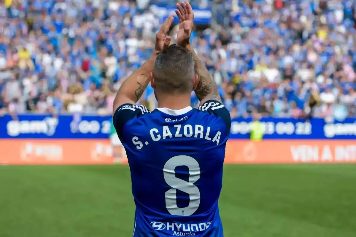 Soccer player wearing the blue number 8 jersey applauding with his back to the crowd in a packed stadium Soccer player wearing the blue number 8 jersey applauding with his back to the crowd in a packed stadium