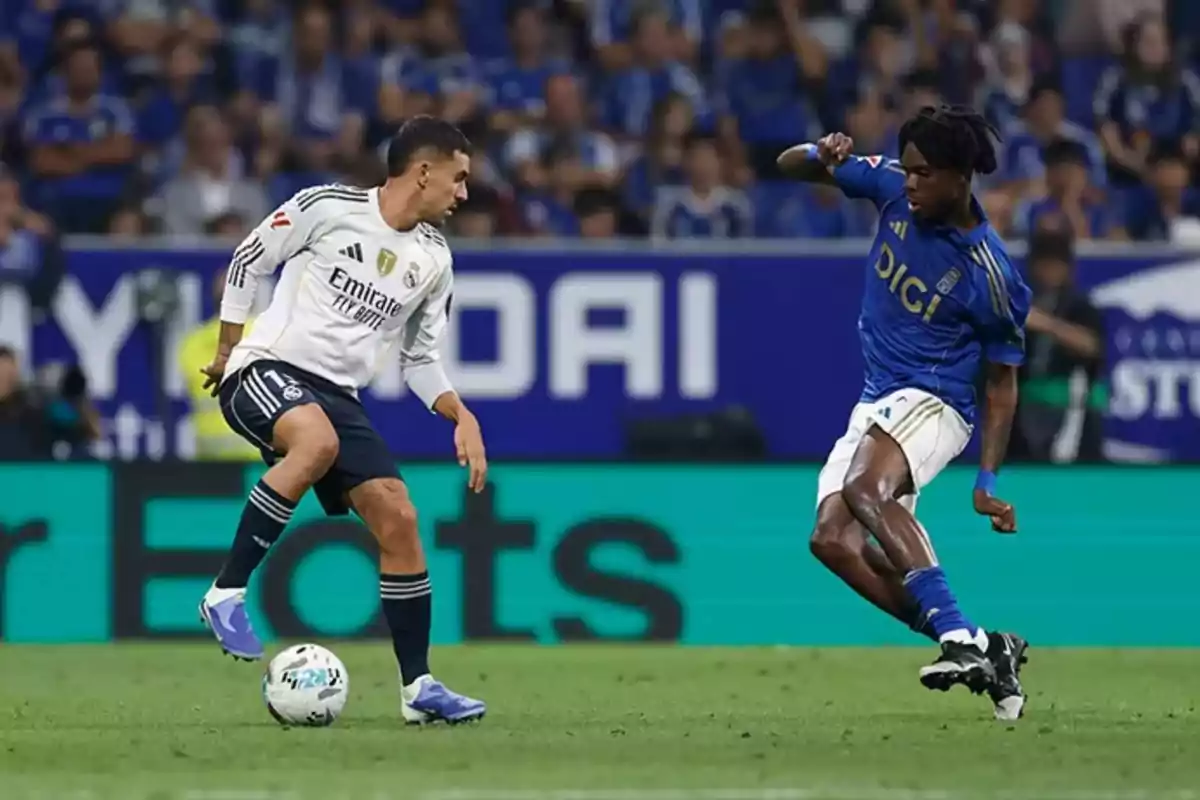 Dos futbolistas disputan el balón durante un partido, uno con uniforme blanco y el otro con uniforme azul, en un estadio lleno de espectadores. Dos futbolistas disputan el balón durante un partido, uno con uniforme blanco y el otro con uniforme azul, en un estadio lleno de espectadores.