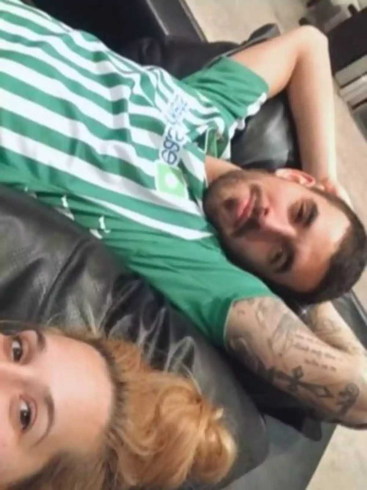 Dos personas recostadas en un sillón negro, una de ellas lleva una camiseta de rayas verdes y blancas y tiene tatuajes en el brazo, la otra persona tiene el cabello rubio recogido. Dos personas recostadas en un sillón negro, una de ellas lleva una camiseta de rayas verdes y blancas y tiene tatuajes en el brazo, la otra persona tiene el cabello rubio recogido.
