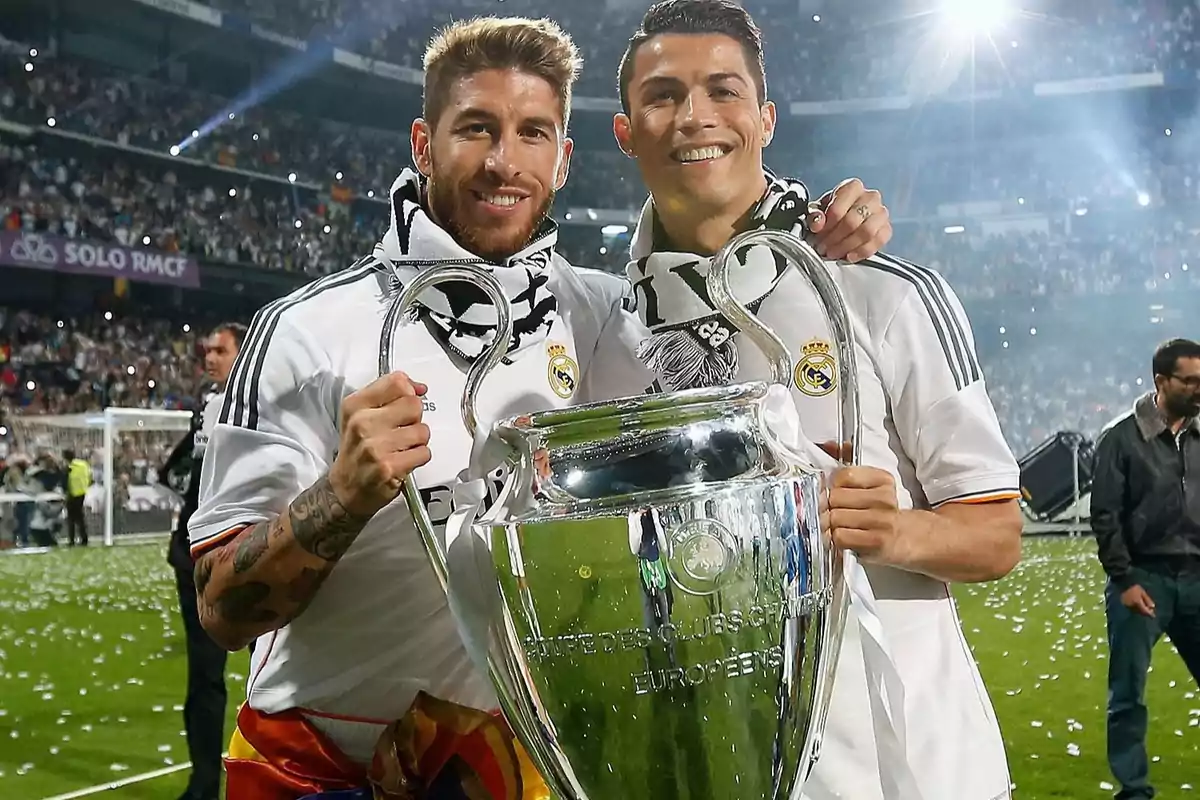Dos futbolistas del Real Madrid sonríen y posan juntos sosteniendo el trofeo de la Champions League en un estadio lleno de aficionados celebrando Dos futbolistas del Real Madrid sonríen y posan juntos sosteniendo el trofeo de la Champions League en un estadio lleno de aficionados celebrando