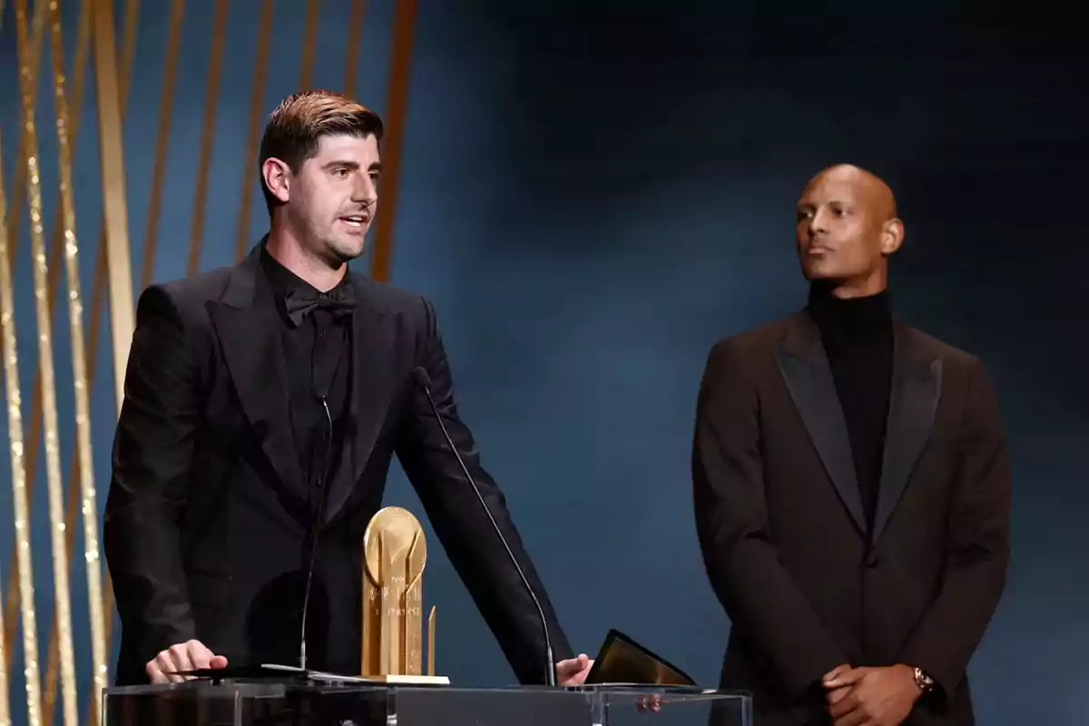 A man in a black tuxedo speaks at a podium next to a golden trophy while another man in a dark suit watches him on the stage of a ceremony. A man in a black tuxedo speaks at a podium next to a golden trophy while another man in a dark suit watches him on the stage of a ceremony.