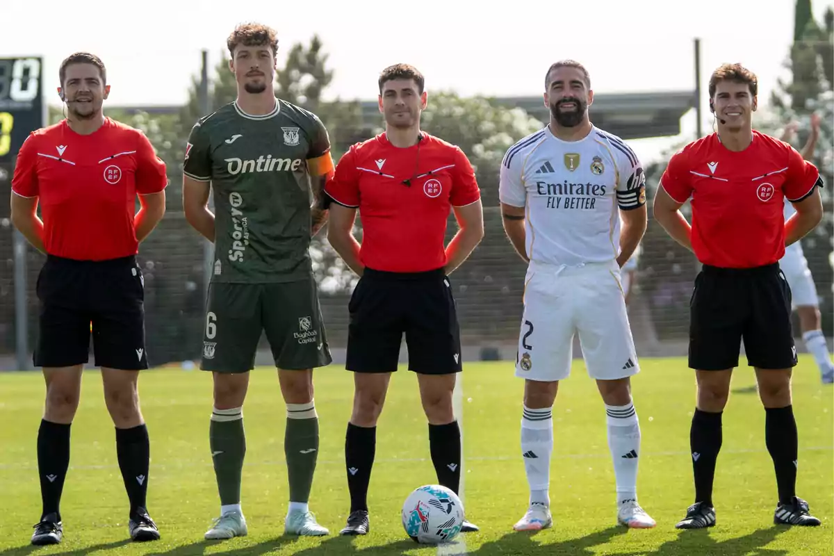 Cinco personas posan en el campo de fútbol, dos jugadores con uniforme y tres árbitros con camiseta roja y pantalón negro, con un balón en el césped frente a ellos Cinco personas posan en el campo de fútbol, dos jugadores con uniforme y tres árbitros con camiseta roja y pantalón negro, con un balón en el césped frente a ellos