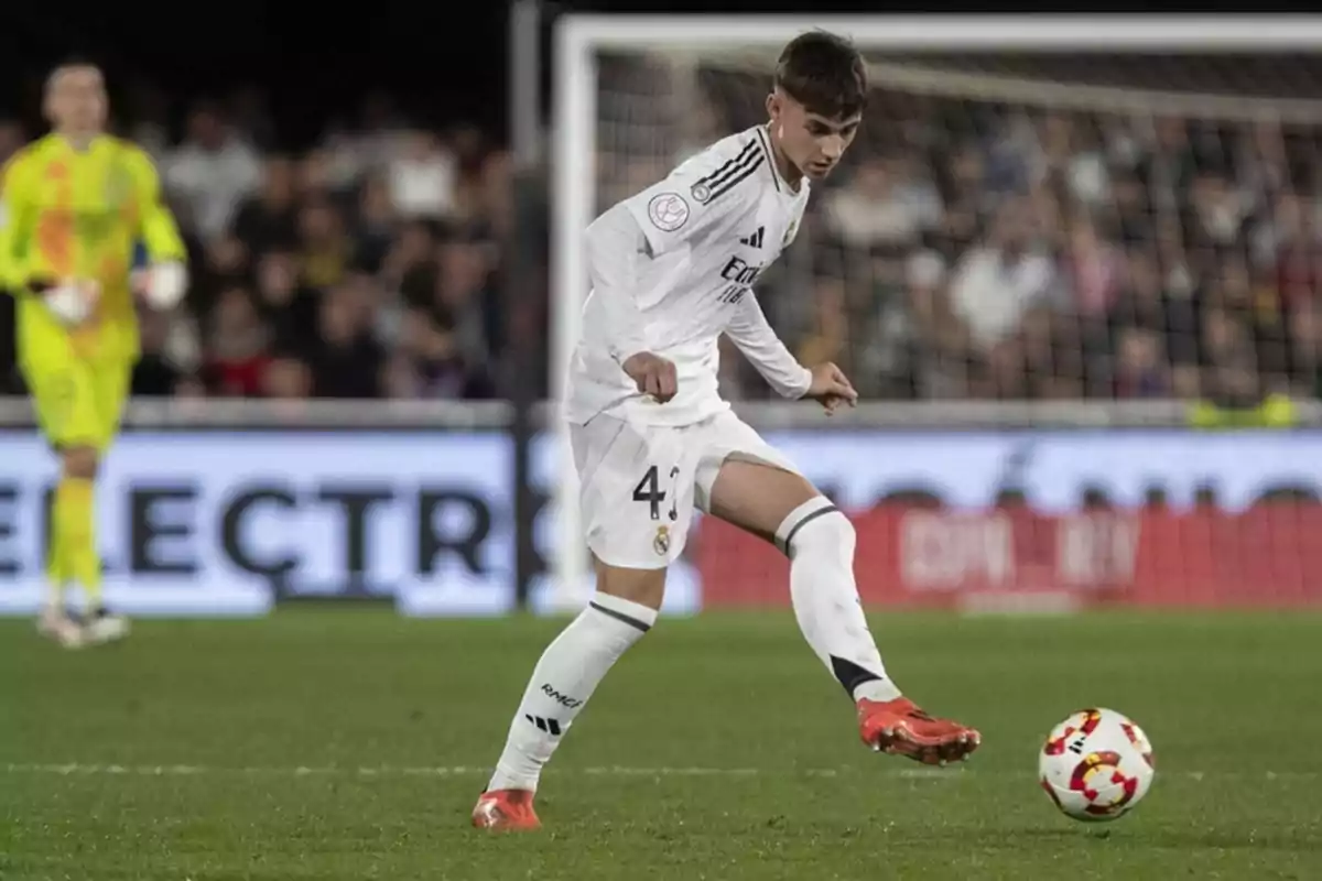 Jugador de fútbol con uniforme blanco controlando el balón en un partido con el portero de fondo y público en las gradas Jugador de fútbol con uniforme blanco controlando el balón en un partido con el portero de fondo y público en las gradas