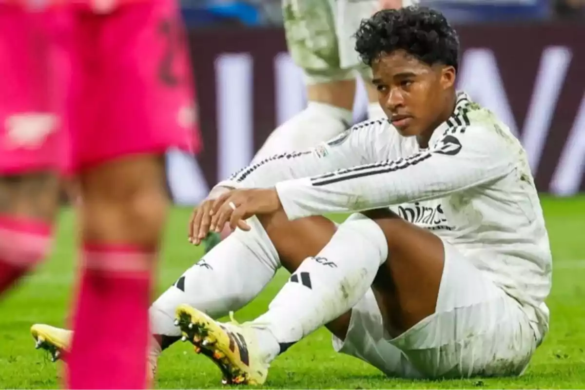 Jugador de fútbol con uniforme blanco sentado en el césped durante un partido mostrando una expresión de cansancio o frustración Jugador de fútbol con uniforme blanco sentado en el césped durante un partido mostrando una expresión de cansancio o frustración