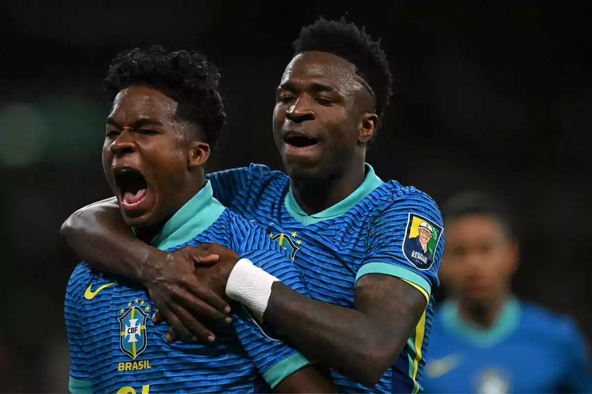 Two players from the Brazil national team celebrate a goal with excitement during a match; one of them shouts while the other hugs him from behind. Two players from the Brazil national team celebrate a goal with excitement during a match; one of them shouts while the other hugs him from behind.