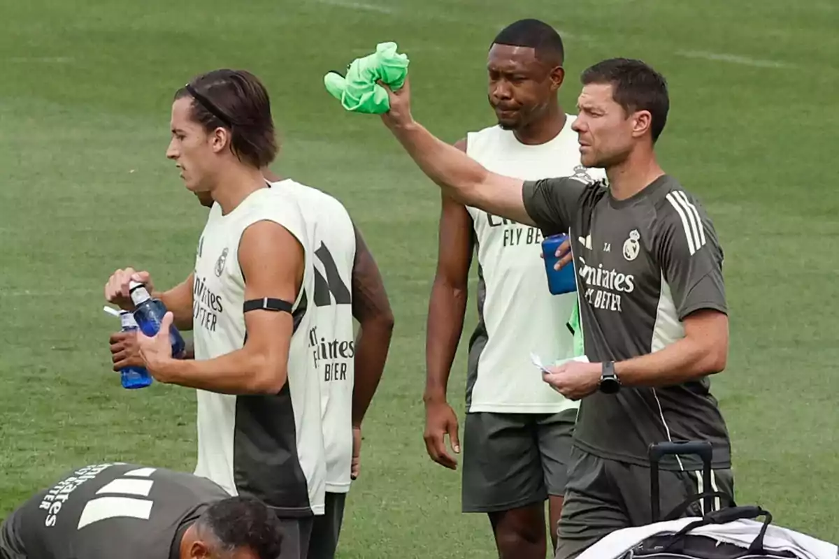 Entrenador y jugadores del Real Madrid durante una sesión de entrenamiento en el campo, uno de ellos sostiene una toalla verde y otro tiene una botella de agua Entrenador y jugadores del Real Madrid durante una sesión de entrenamiento en el campo, uno de ellos sostiene una toalla verde y otro tiene una botella de agua