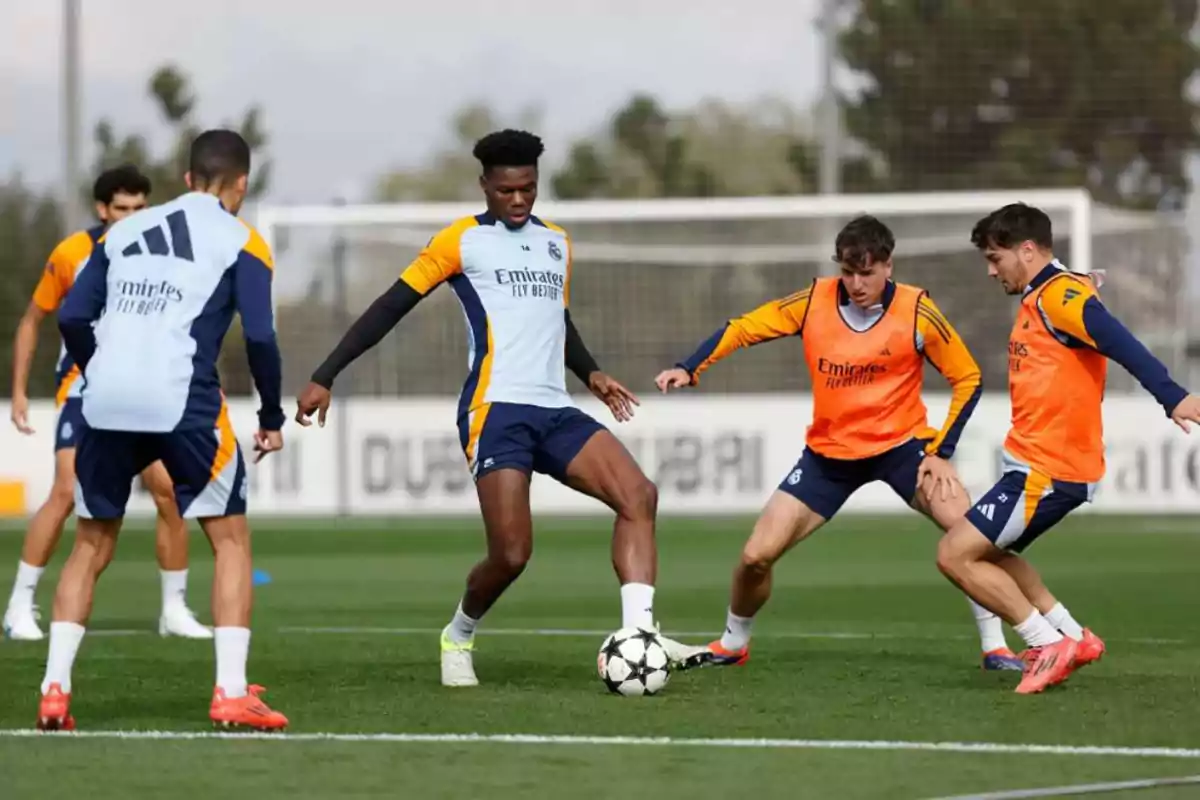 Jugadores de fútbol entrenando en un campo con petos naranjas y uniformes de entrenamiento azul y amarillo mientras disputan el balón Jugadores de fútbol entrenando en un campo con petos naranjas y uniformes de entrenamiento azul y amarillo mientras disputan el balón