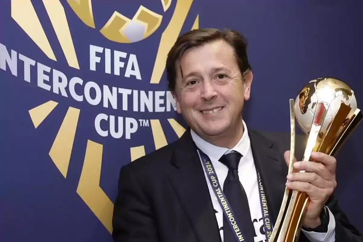 Smiling man in a suit holding a golden trophy in front of a background with the FIFA Intercontinental Cup logo Smiling man in a suit holding a golden trophy in front of a background with the FIFA Intercontinental Cup logo