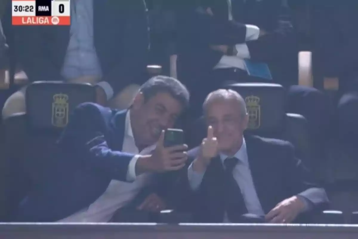 Dos hombres con traje se toman una selfie en un estadio mientras uno de ellos sonríe y muestra el pulgar arriba