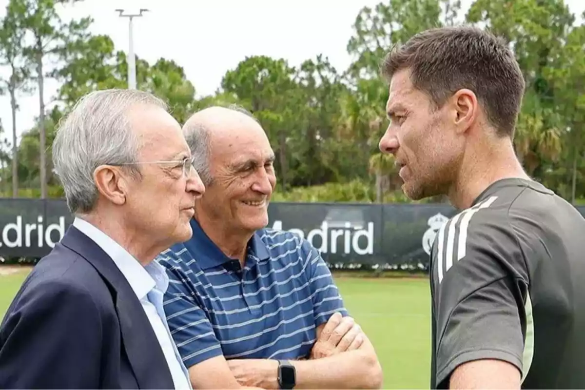 Tres hombres conversan en un campo de entrenamiento con árboles al fondo y un cartel de Madrid parcialmente visible. Tres hombres conversan en un campo de entrenamiento con árboles al fondo y un cartel de Madrid parcialmente visible.