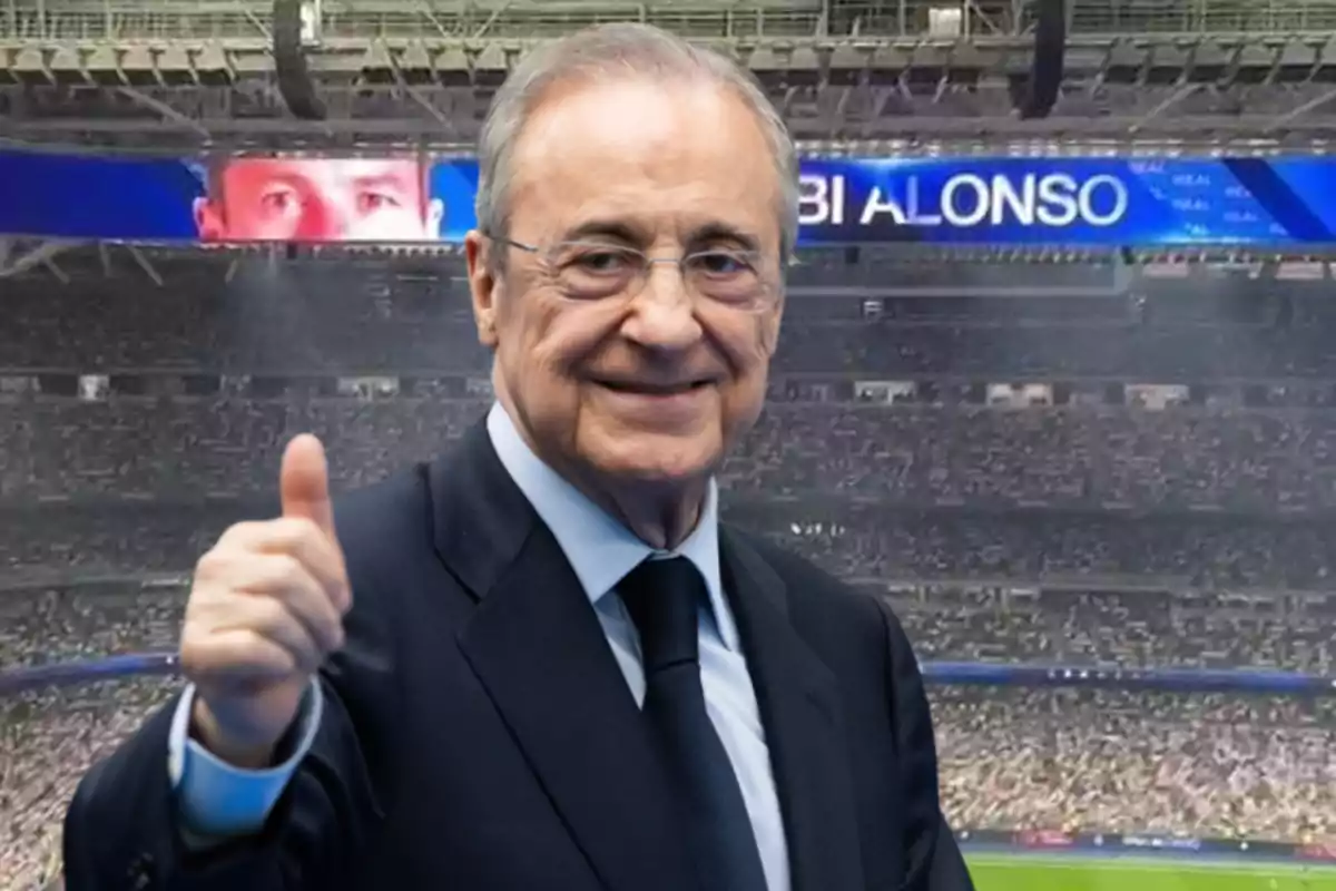 Smiling man in a suit giving a thumbs up in a stadium full of spectators with a giant screen in the background