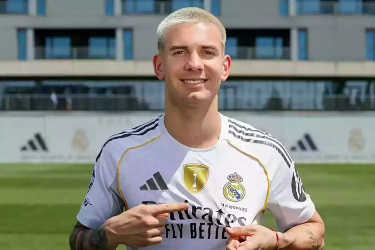 Futbolista con cabello rubio corto sonríe y señala el escudo del Real Madrid en su camiseta blanca durante el día en un campo de entrenamiento Futbolista con cabello rubio corto sonríe y señala el escudo del Real Madrid en su camiseta blanca durante el día en un campo de entrenamiento
