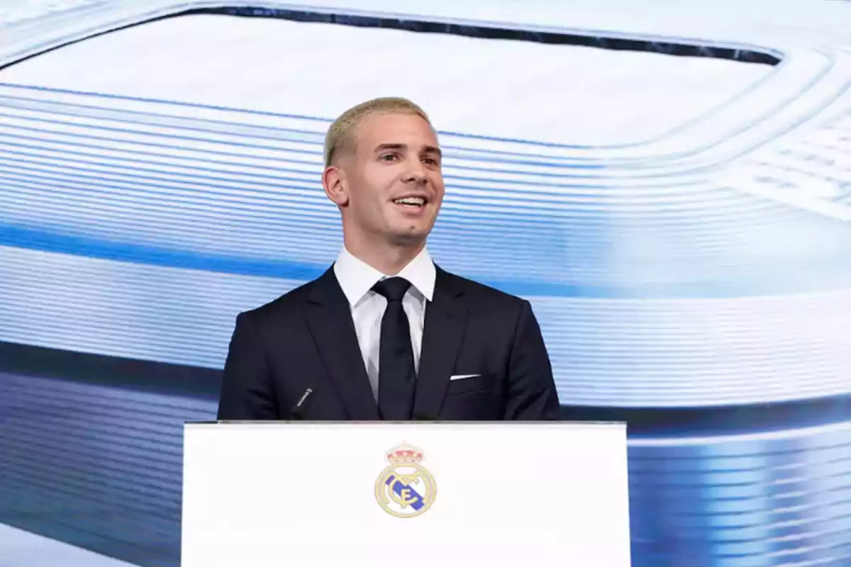 A man with short blond hair, dressed in a dark suit and black tie, smiles while speaking at a podium with the Real Madrid crest on the front and a light blue background. A man with short blond hair, dressed in a dark suit and black tie, smiles while speaking at a podium with the Real Madrid crest on the front and a light blue background.
