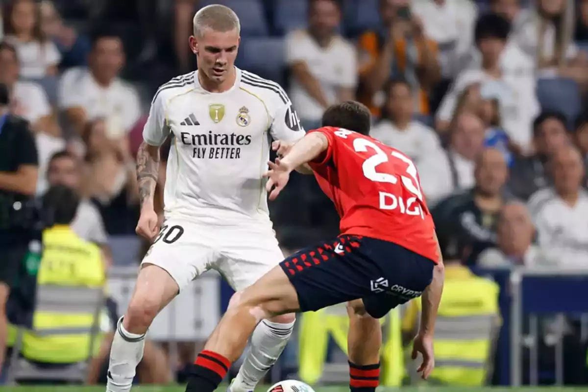 Two players compete for the ball during a match, one wearing a white uniform and the other wearing a red uniform, while the crowd watches from the stands. Two players compete for the ball during a match, one wearing a white uniform and the other wearing a red uniform, while the crowd watches from the stands.