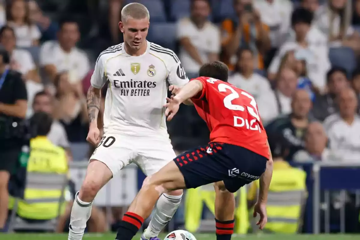 Two players compete for the ball during a match, one wearing a white uniform and the other wearing a red uniform, while the crowd watches from the stands. Two players compete for the ball during a match, one wearing a white uniform and the other wearing a red uniform, while the crowd watches from the stands.